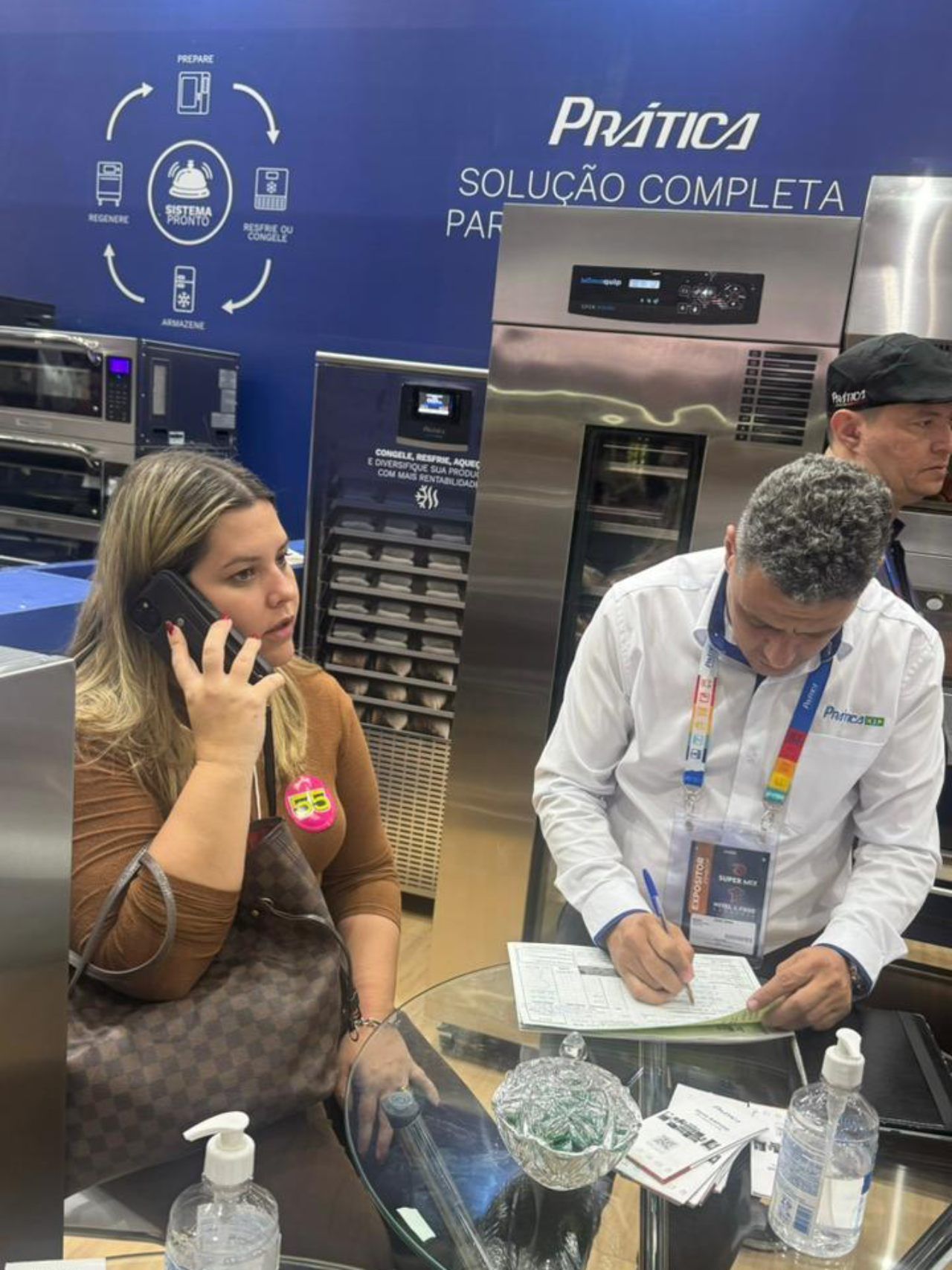 Uma mulher está falando ao celular enquanto um homem escreve em um pedaço de papel.