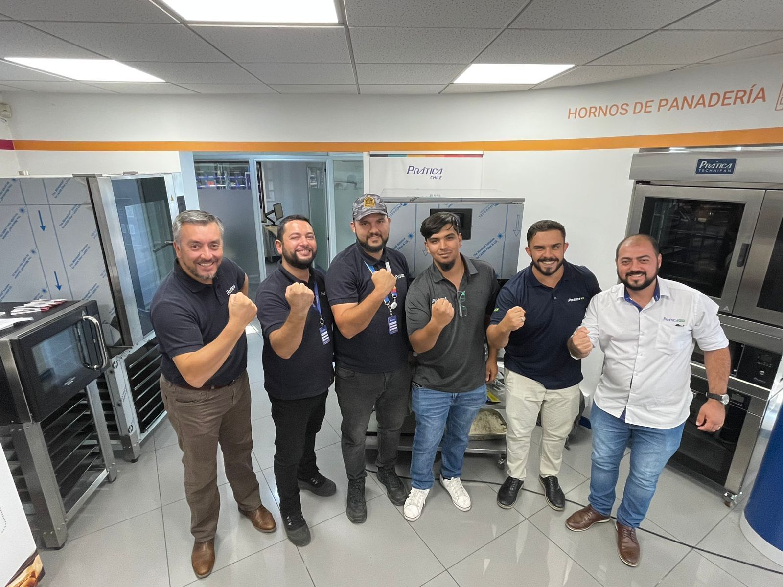 Seis homens em uma padaria posando com os punhos cerrados. Fornos de aço inoxidável alinham-se nas paredes.