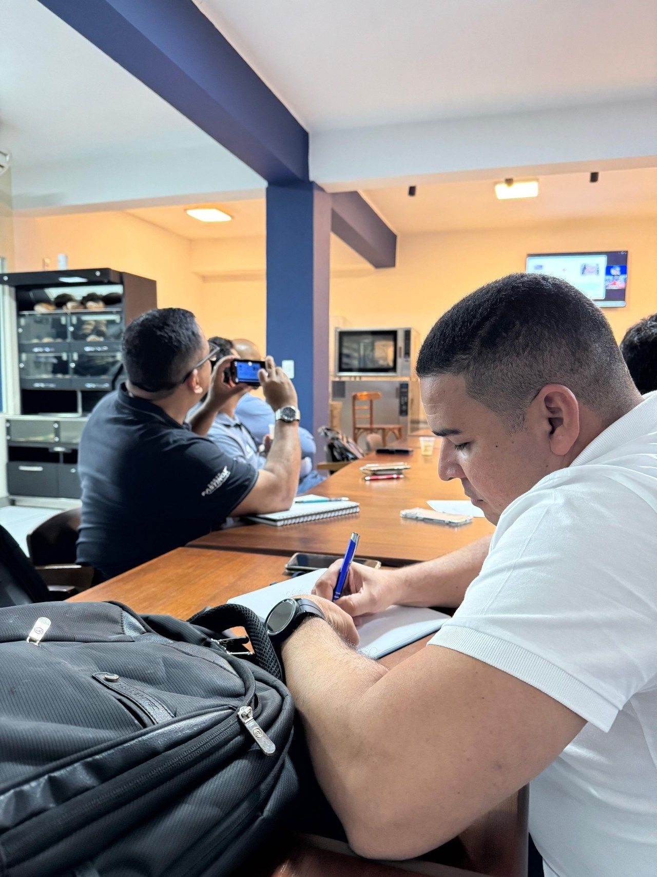 Pessoas sentadas à mesa, fazendo anotações e usando smartphones, em uma sala com uma janela e uma grande viga.