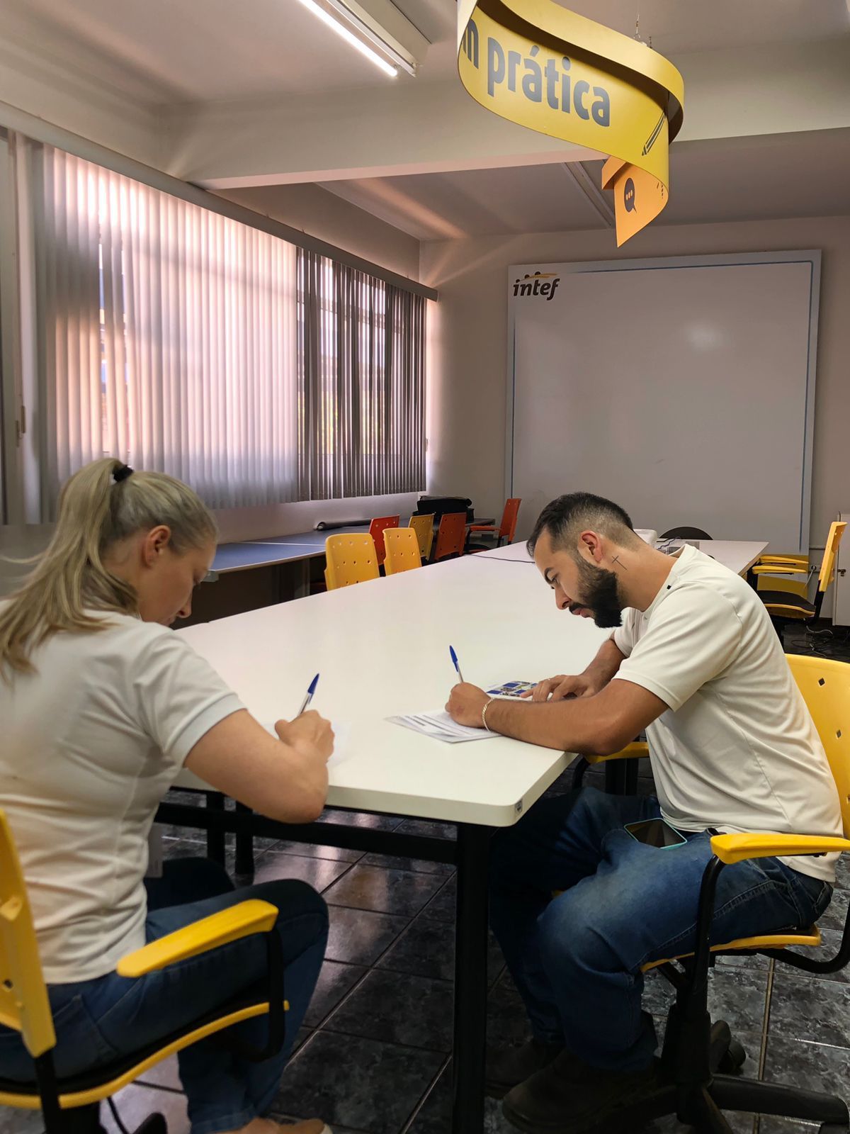 Um homem e uma mulher estão sentados em uma longa mesa escrevendo