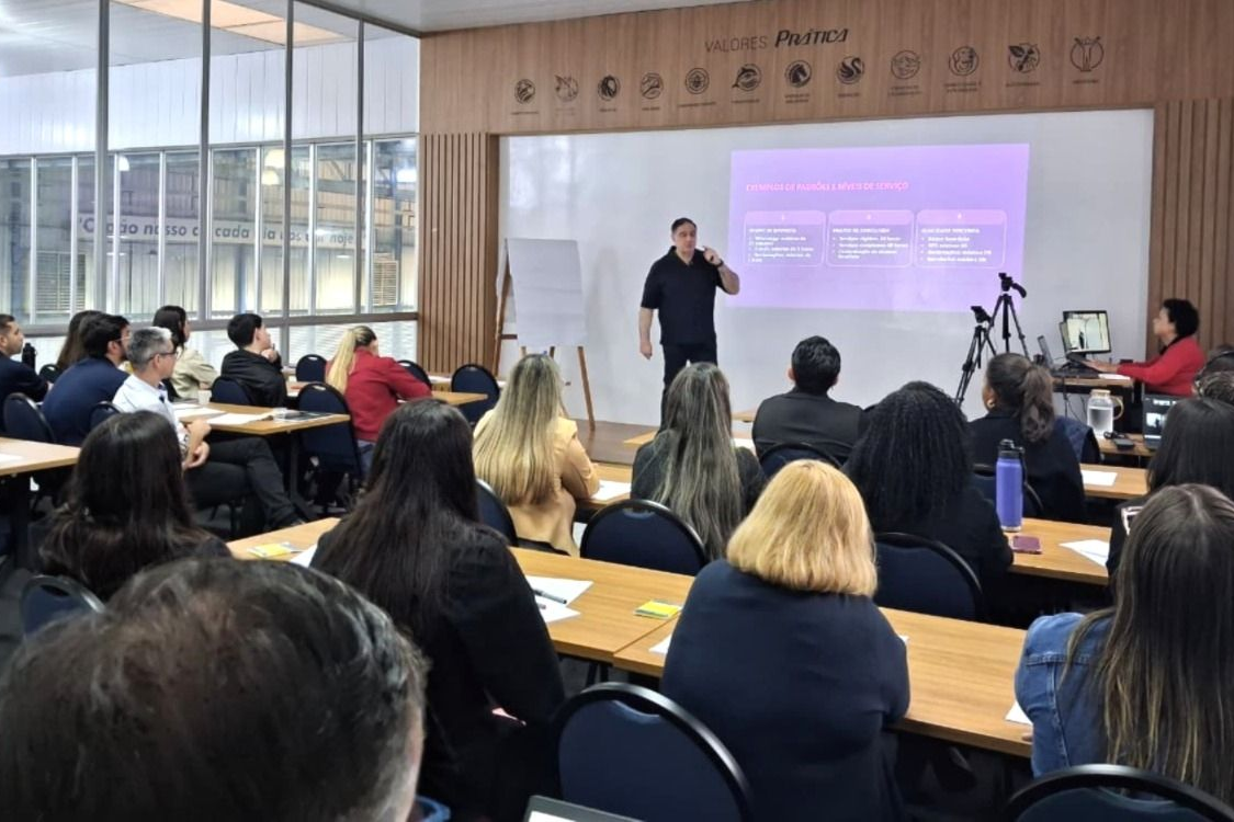 Um homem fazendo uma apresentação para uma sala de aula cheia de pessoas. Tela de projetor e carteiras presentes.
