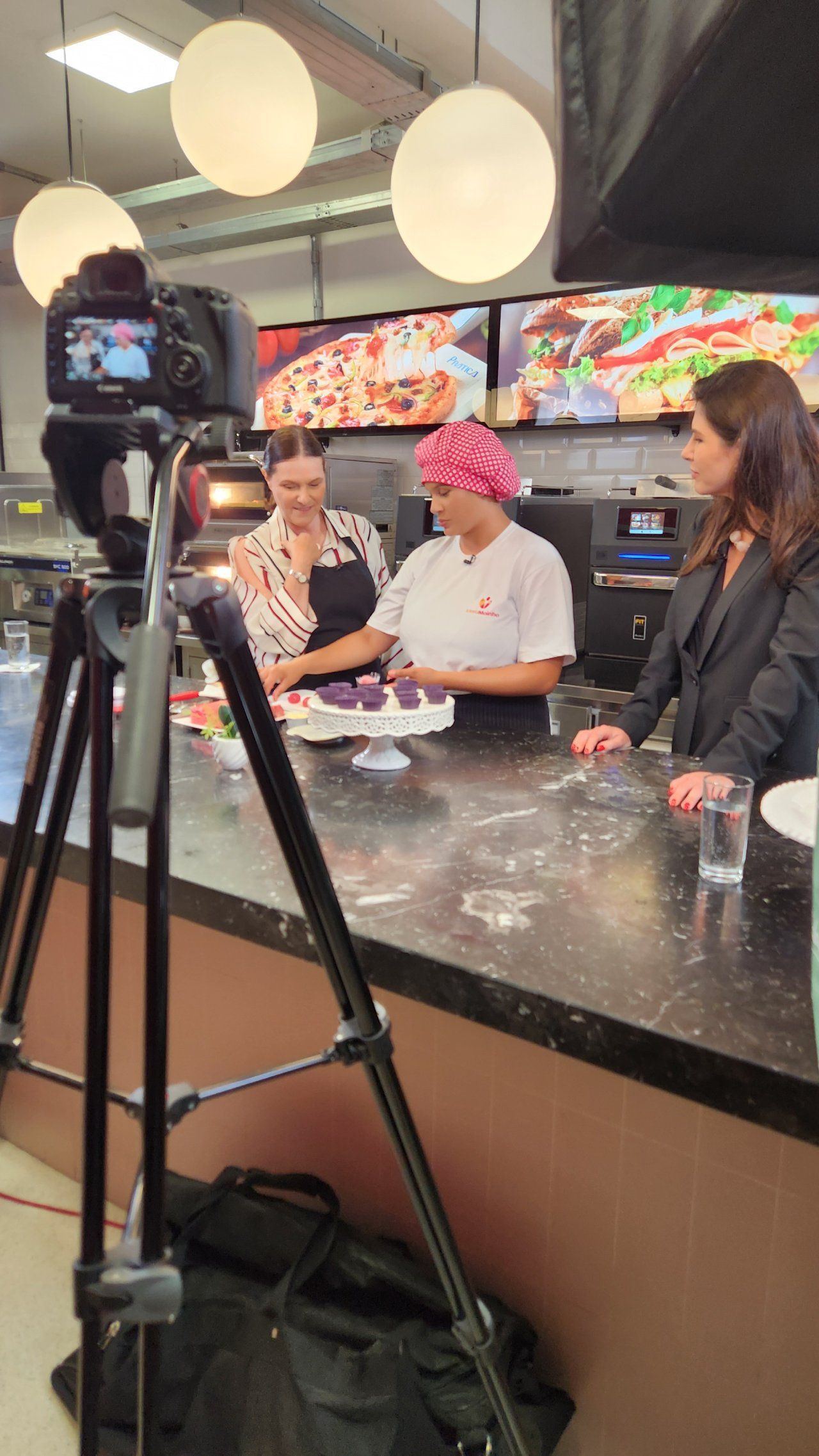 Uma mulher está parada em frente a uma câmera em uma cozinha.