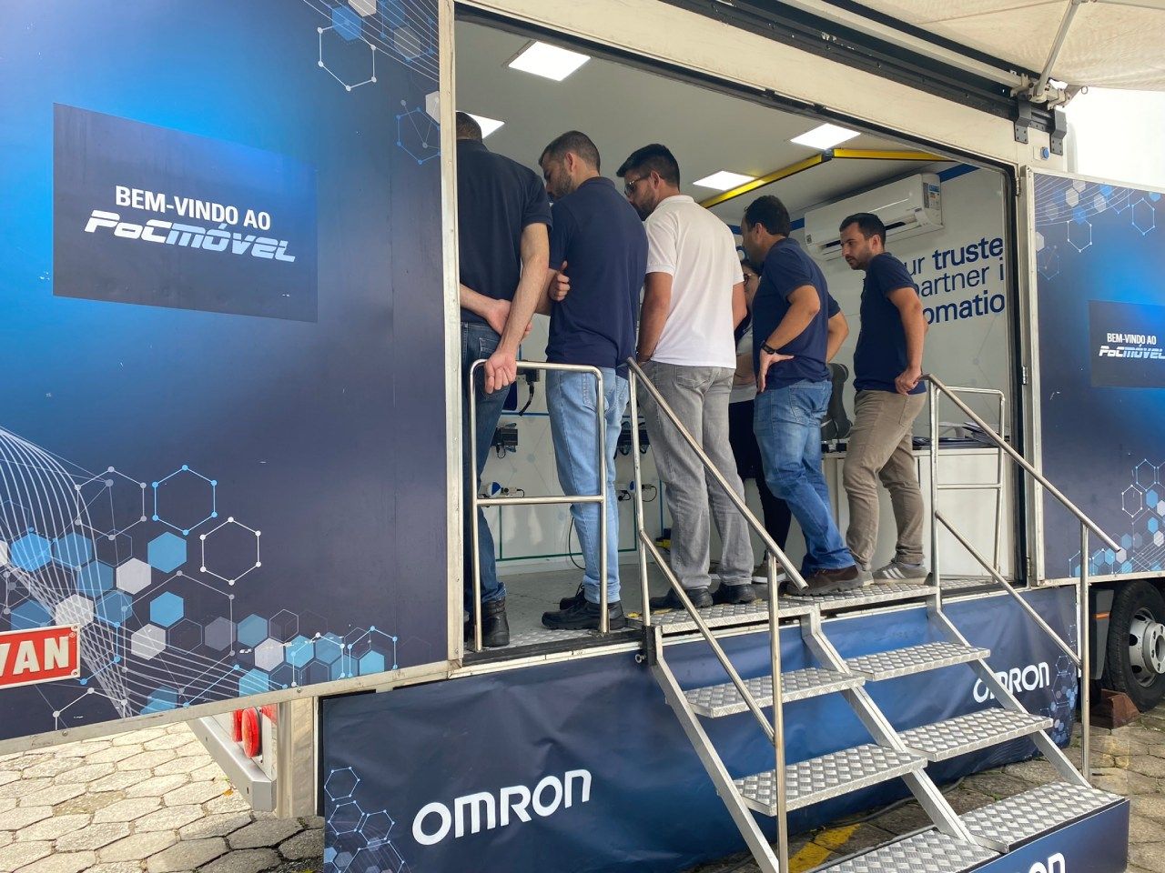 Um grupo de homens está parado em frente a um trailer com a palavra Omron escrita nele.