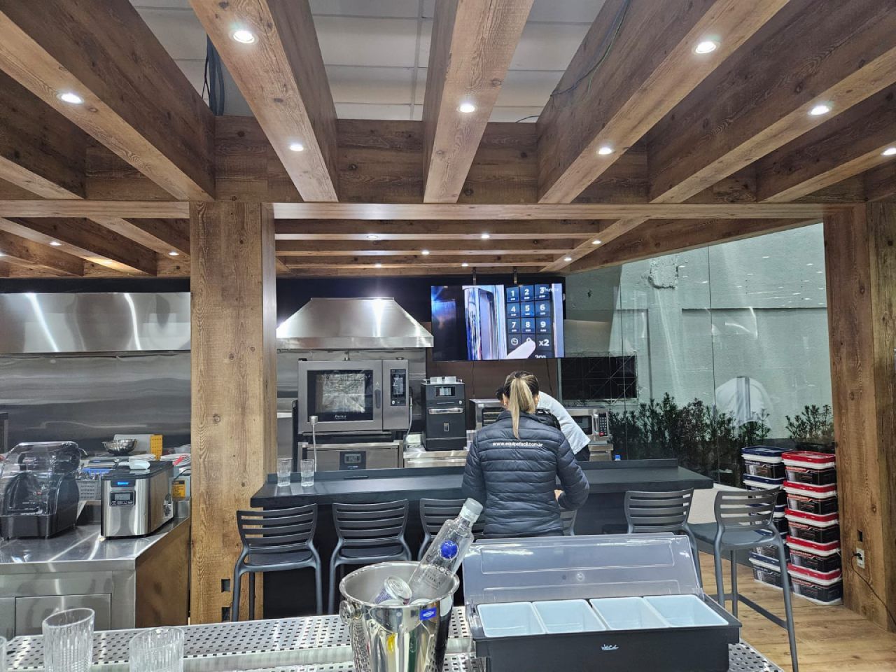 Uma mulher está de pé em uma cozinha com vigas de madeira e eletrodomésticos de aço inoxidável.