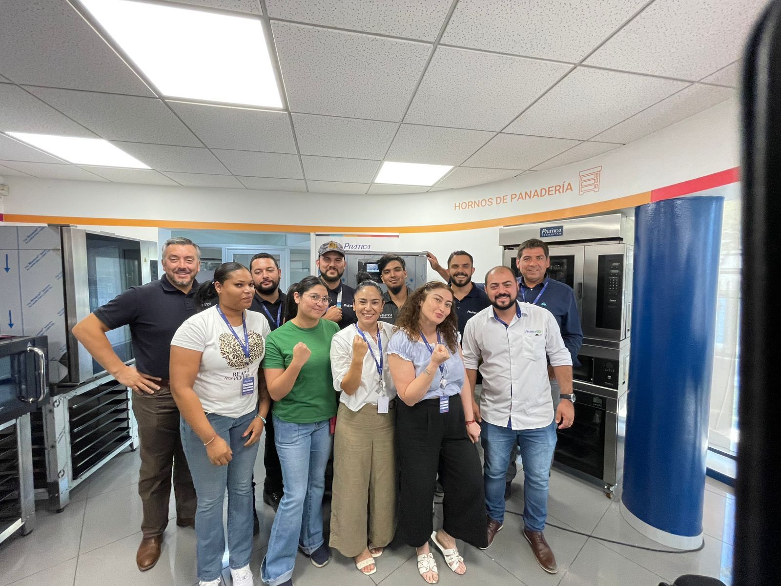 Grupo de pessoas sorrindo, posando em uma cozinha comercial com fornos e eletrodomésticos de aço inoxidável.