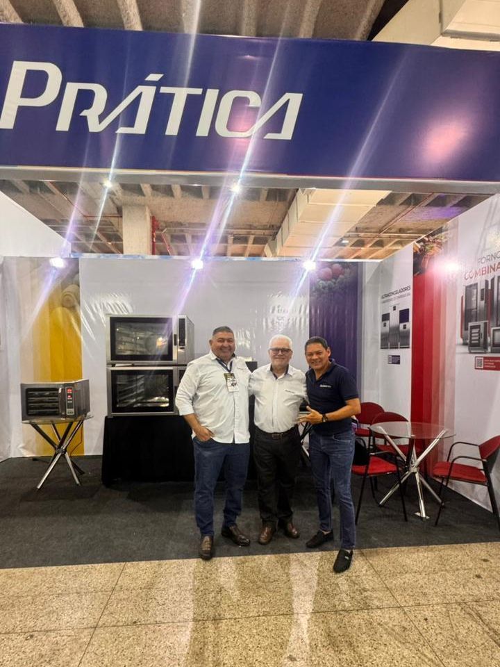 Três homens posam em frente a um estande da Prática em uma feira comercial, com fornos expostos.