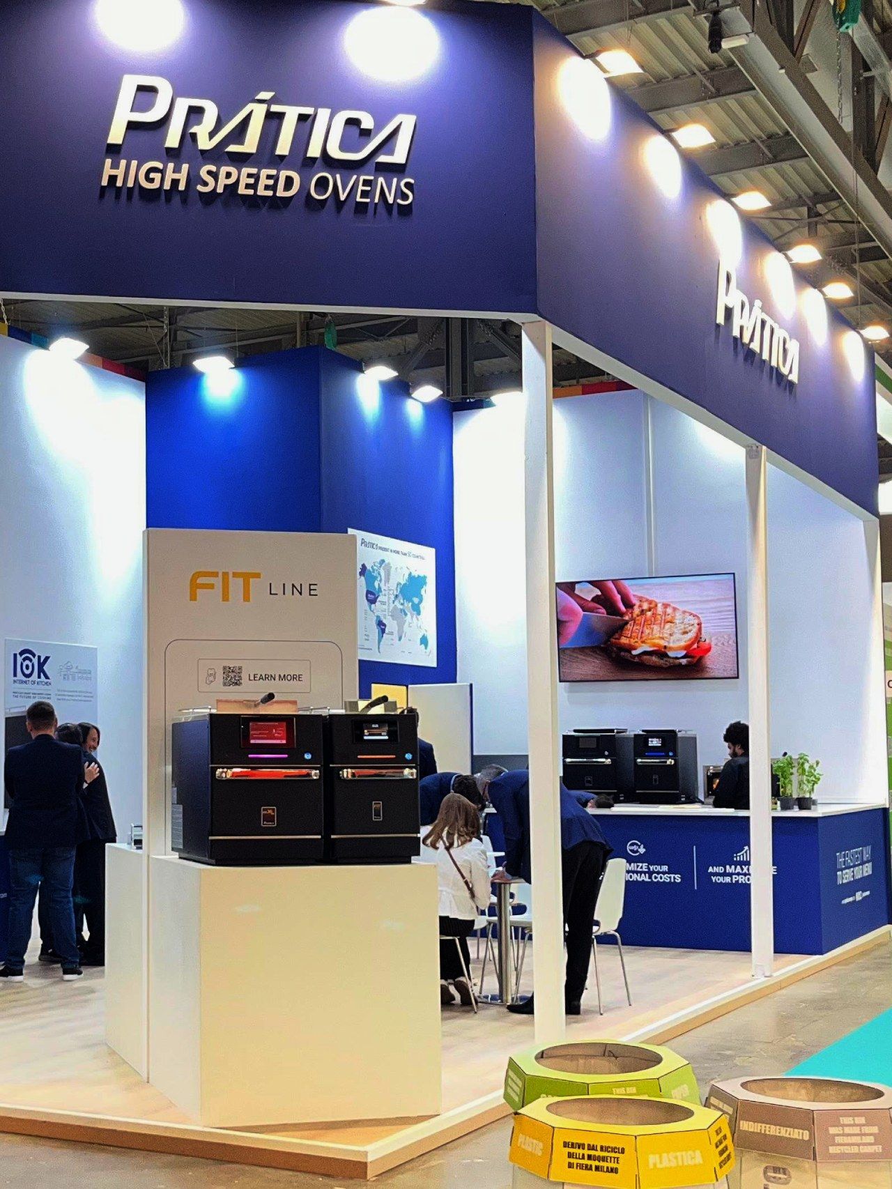 Estande da Prática High Speed ​​Ovens em feira, azul e branco, com fornos em exposição. Pessoas em pé ao redor.