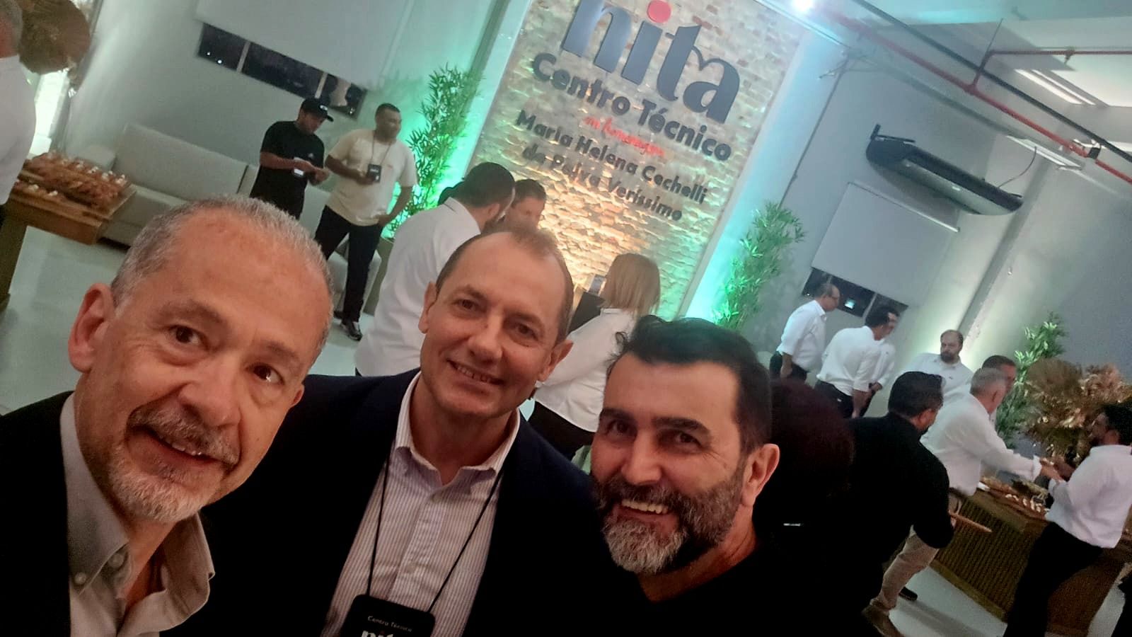 Três homens sorrindo para uma selfie em um evento empresarial, outras pessoas ao fundo.