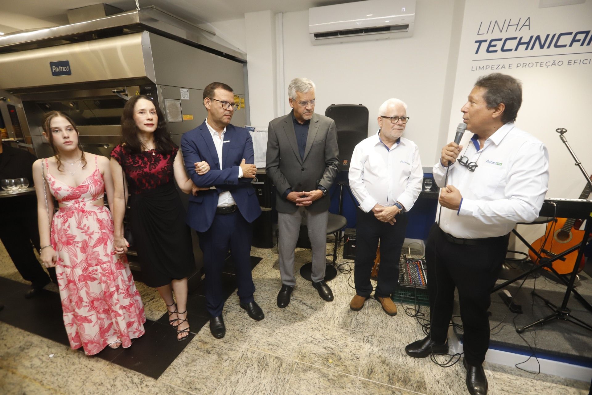 Grupo de pessoas em um evento. Um homem fala ao microfone. Outros ouvem, alguns sorrindo. Ambiente interno.
