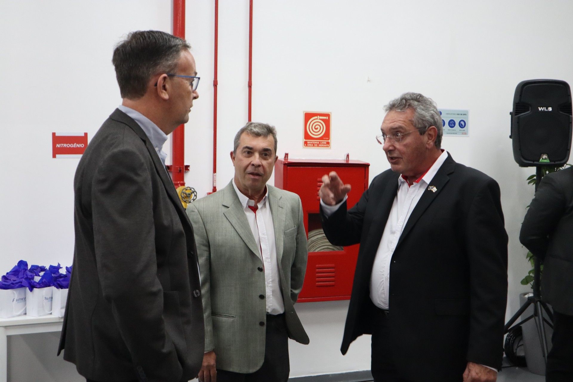 Três homens de terno conversando perto de um armário vermelho para mangueiras de incêndio. Um deles gesticula com a mão. Parede branca.
