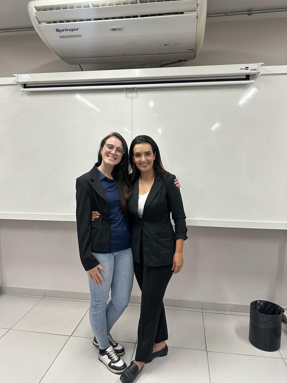 Duas mulheres estão posando para uma foto em frente a um quadro branco.