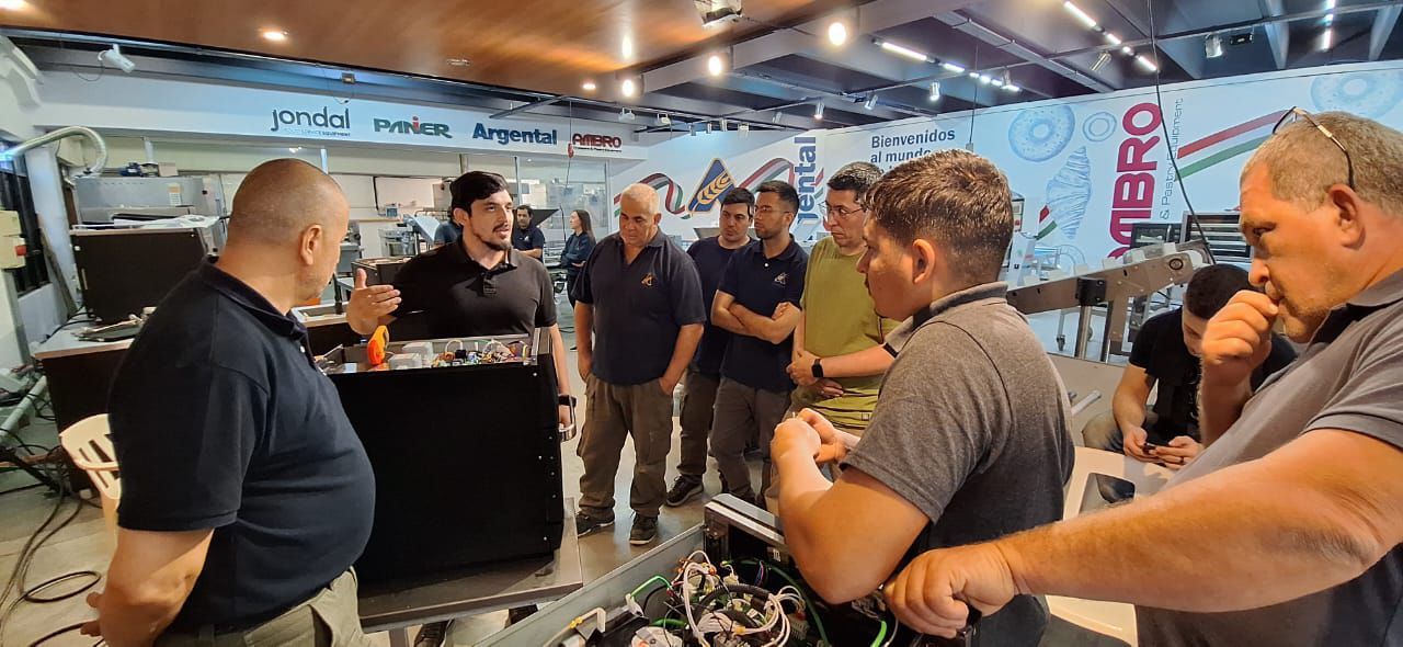 Um grupo de homens em uma oficina, reunidos em volta de uma máquina, parecendo estar observando uma demonstração ou treinamento.