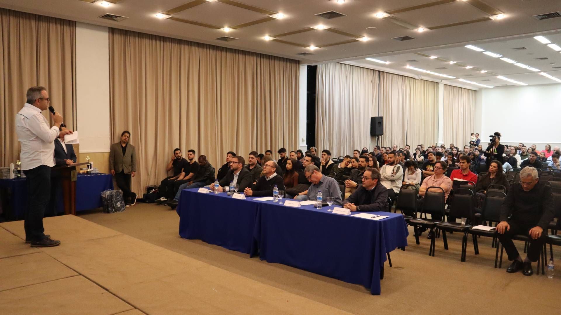 Um homem está de pé na frente de um grande grupo de pessoas sentadas em mesas em uma sala de conferências.