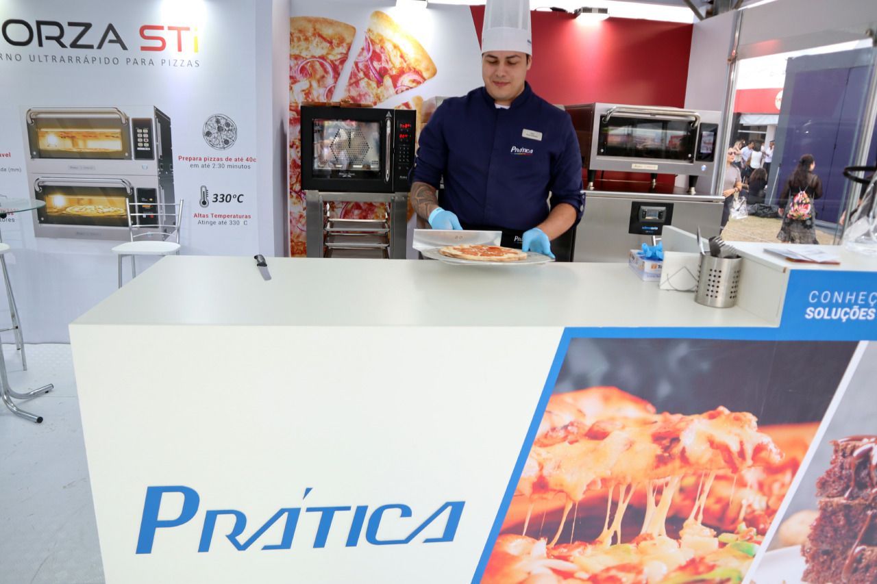 Um homem prepara uma pizza em frente a uma placa que diz pratica