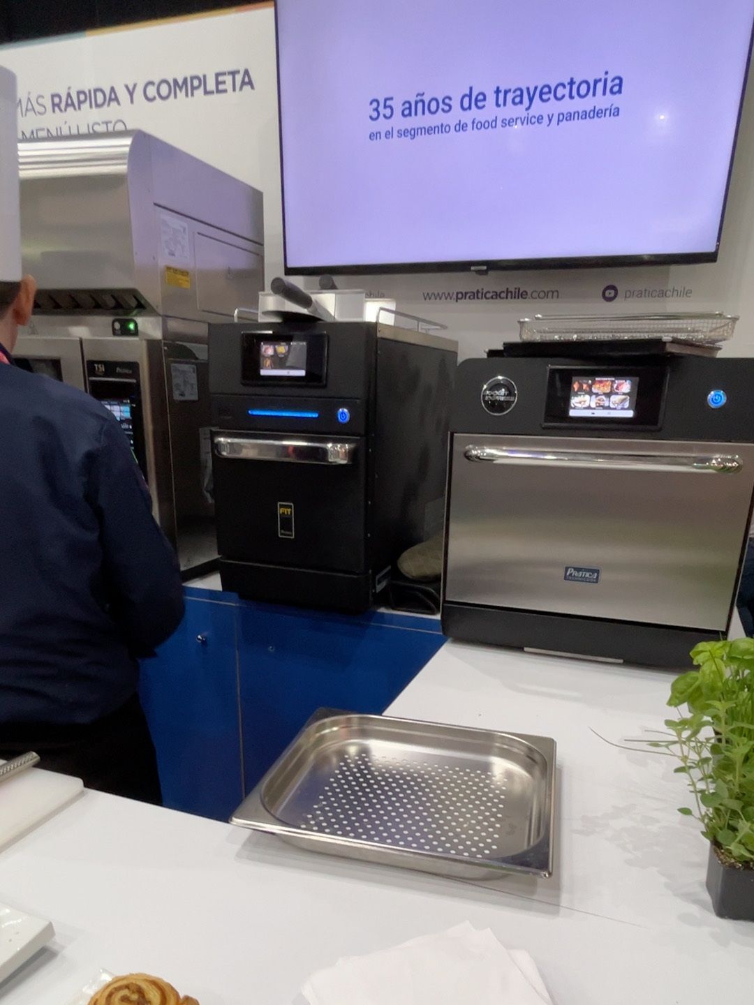 Chef em uma feira de equipamentos alimentícios, demonstrando dois fornos pretos e de aço inoxidável em um balcão.