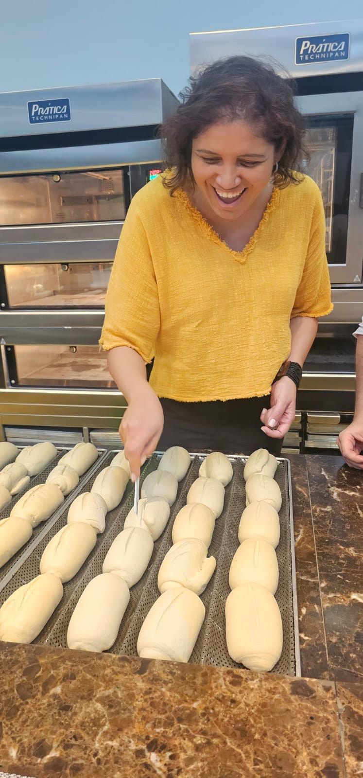 Uma mulher de camisa amarela está parada em frente a uma bandeja de massa.