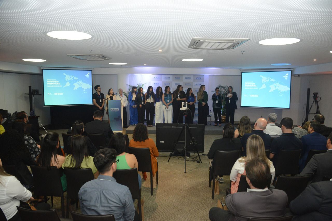 Um grupo de pessoas está sentado em cadeiras em frente a uma tela grande em uma sala de conferências.