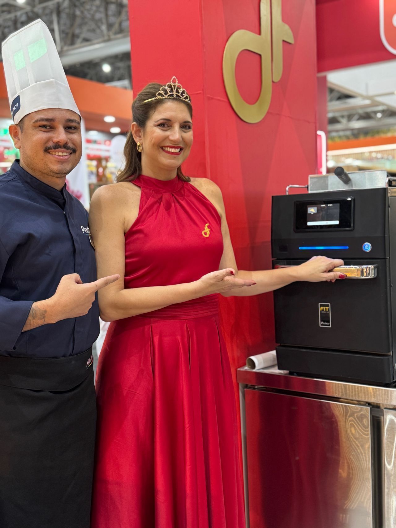 Chef e mulher de vestido vermelho com tiara apontam para uma máquina preta, provavelmente um eletrodoméstico, em um evento.