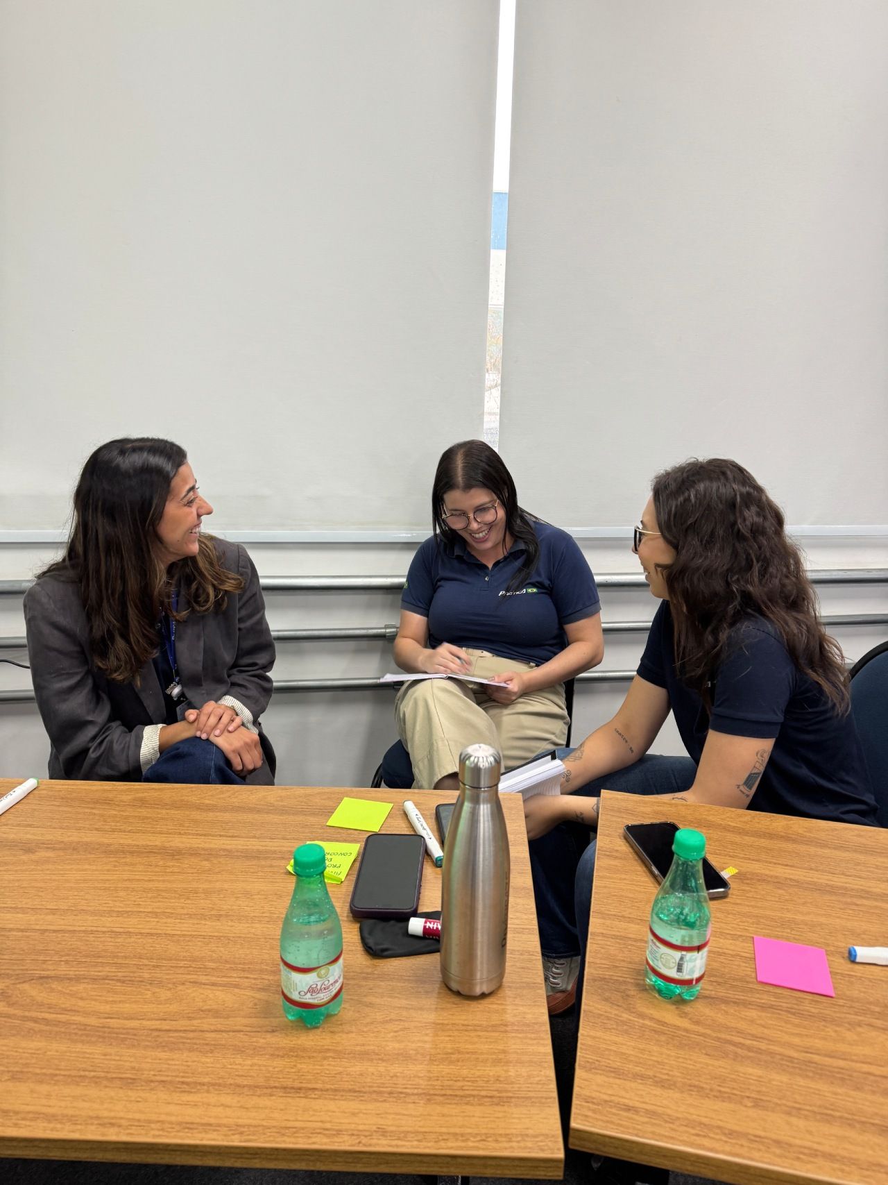 Três pessoas estão sentadas ao redor de mesas de madeira em uma sala, rindo e conversando durante uma reunião colaborativa.