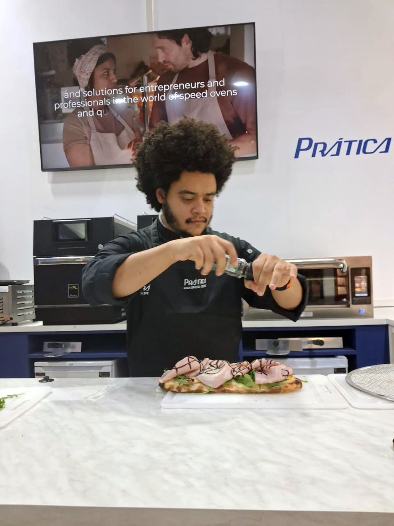 Chef adicionando tempero a um sanduíche. Demonstração culinária com equipamentos de cozinha e uma tela de TV.