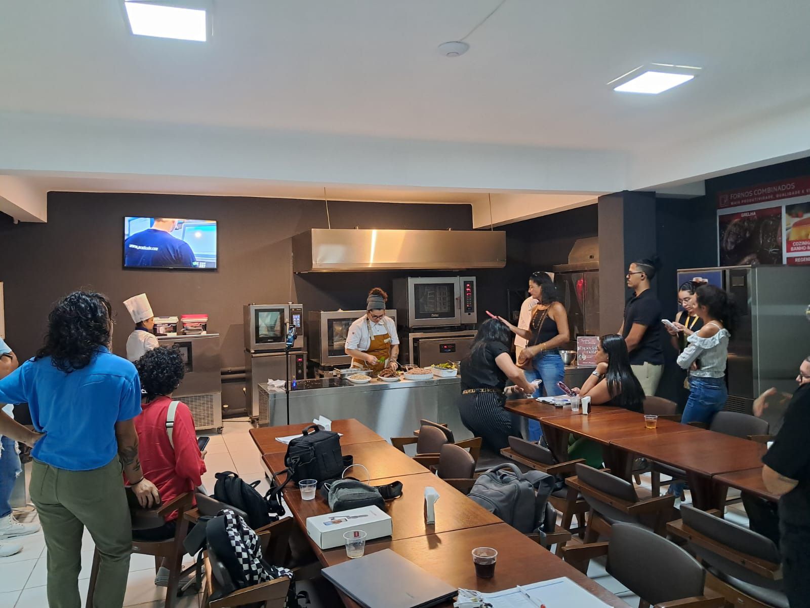 Um grupo de pessoas está de pé ao redor de mesas em uma cozinha.