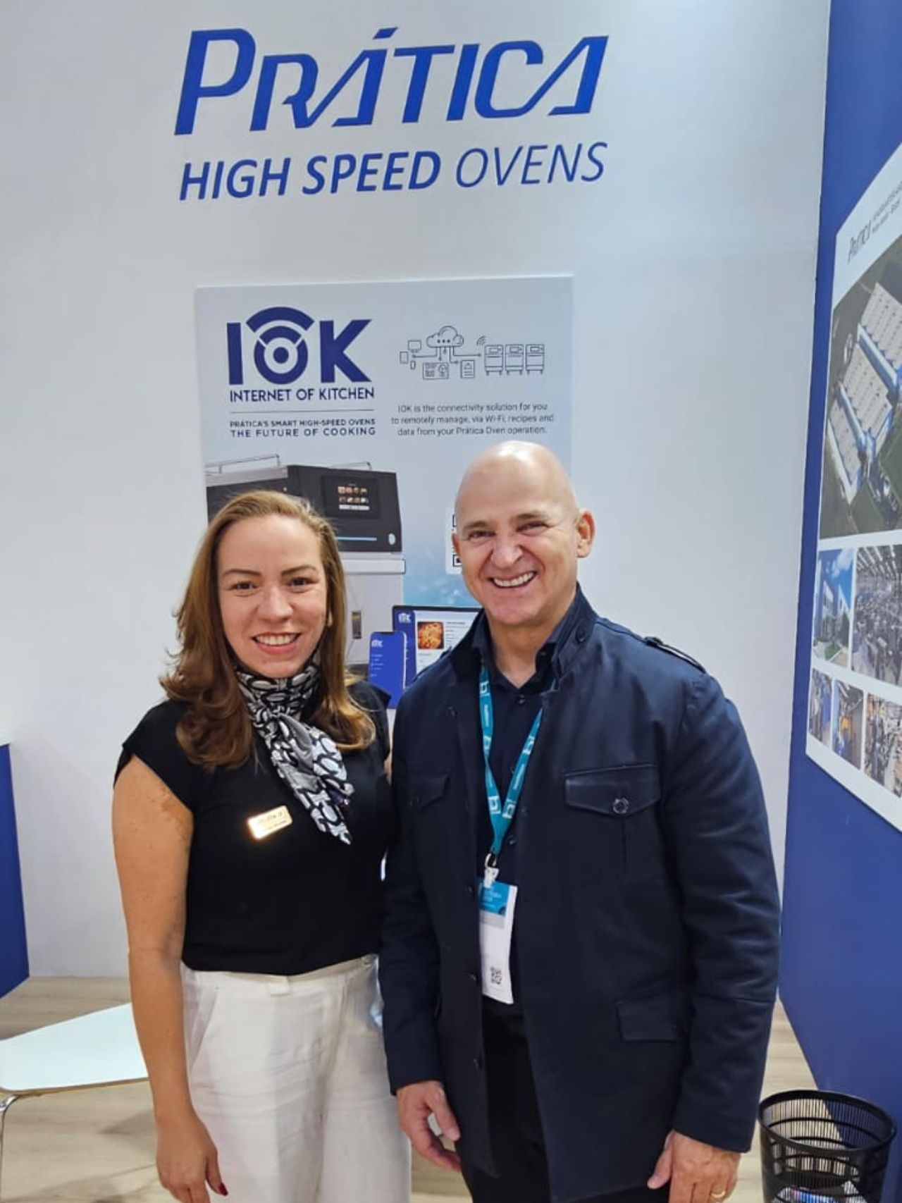 Duas pessoas em frente a um estande da Prática High Speed ​​Ovens. Uma mulher de blusa preta e calça branca está ao lado de um homem de jaqueta azul-marinho.