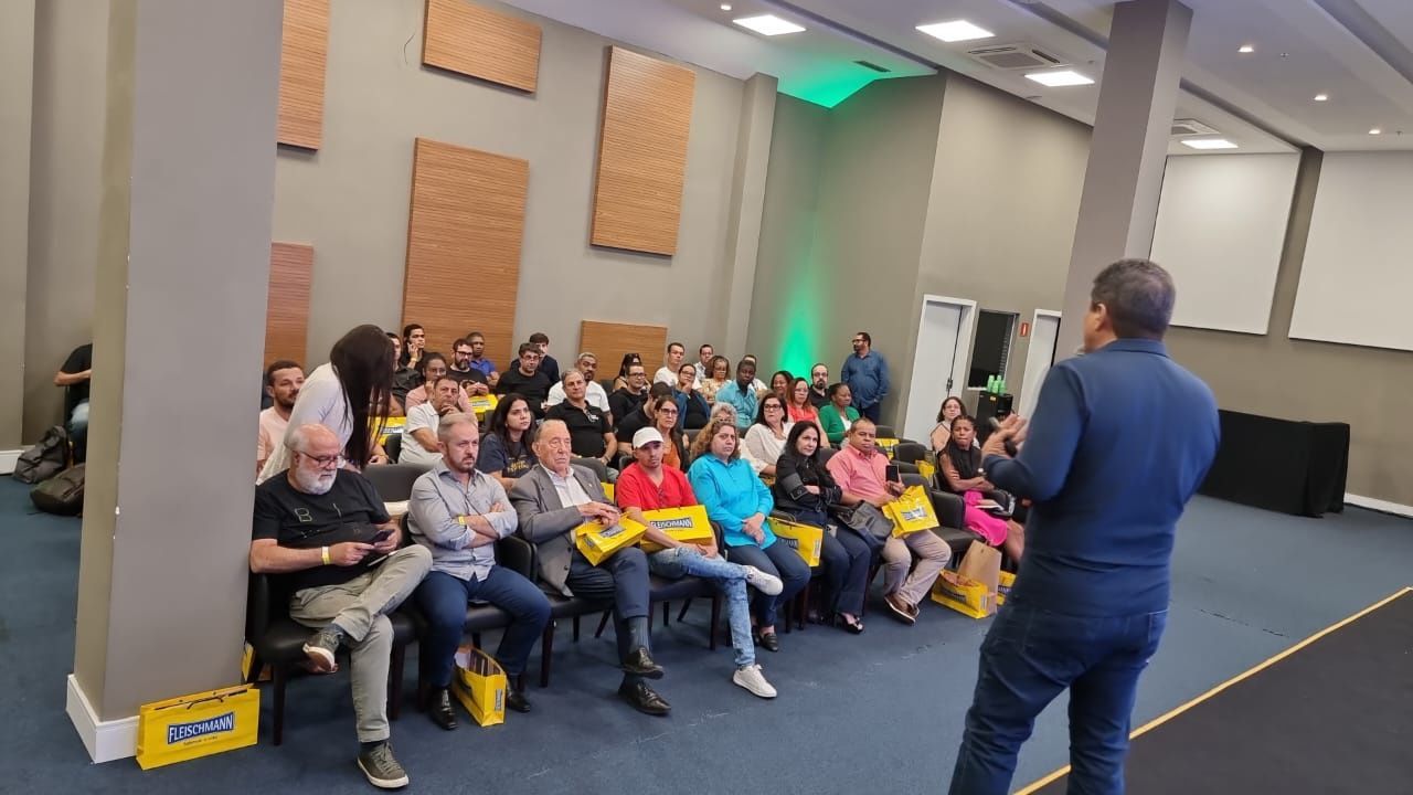 Um homem está de pé na frente de um grande grupo de pessoas sentadas em cadeiras.