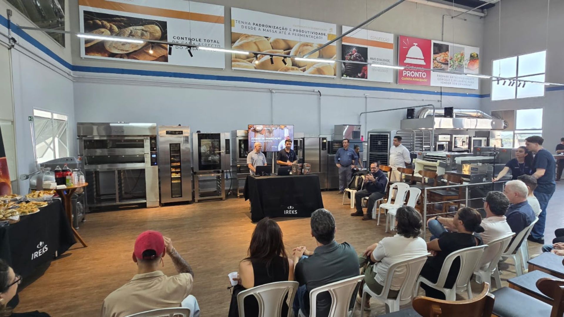 Oficina de panificação: Chefs demonstrando para uma plateia sentada. Cozinha industrial com fornos, doces e banners.