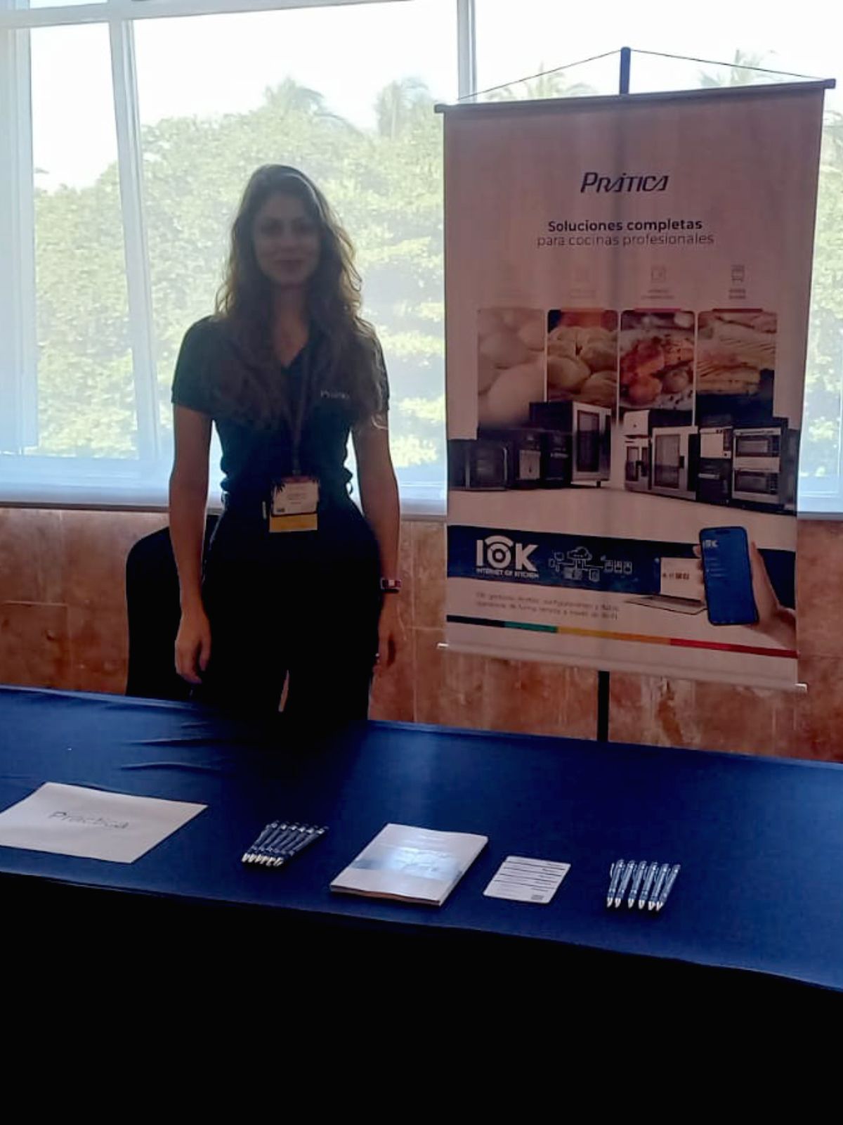 Mulher em um estande de feira comercial com um banner de produto, sorrindo, em uma mesa com papéis e canetas.