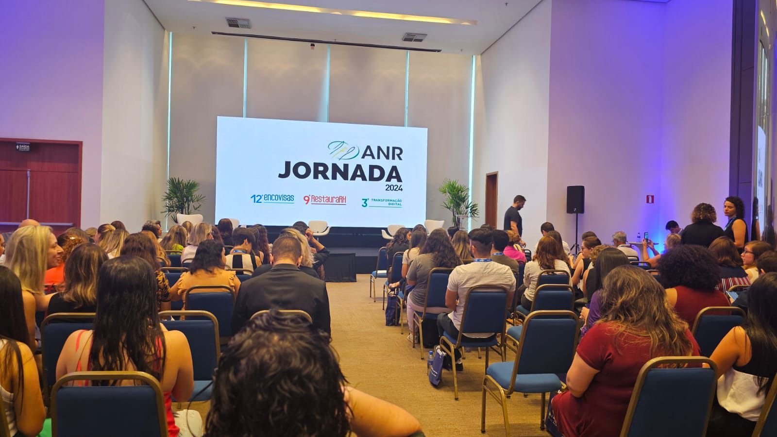 Um grande grupo de pessoas está sentado em cadeiras em frente a uma tela grande que diz jornada.