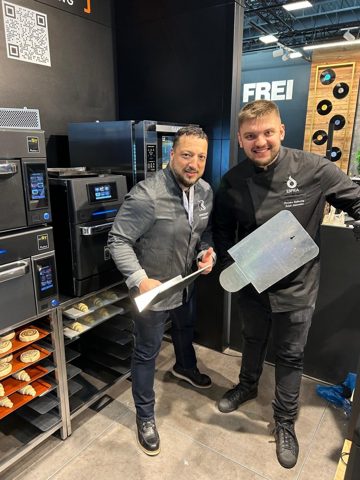 Dois homens estão de pé, um ao lado do outro, em uma cozinha, segurando uma espátula.