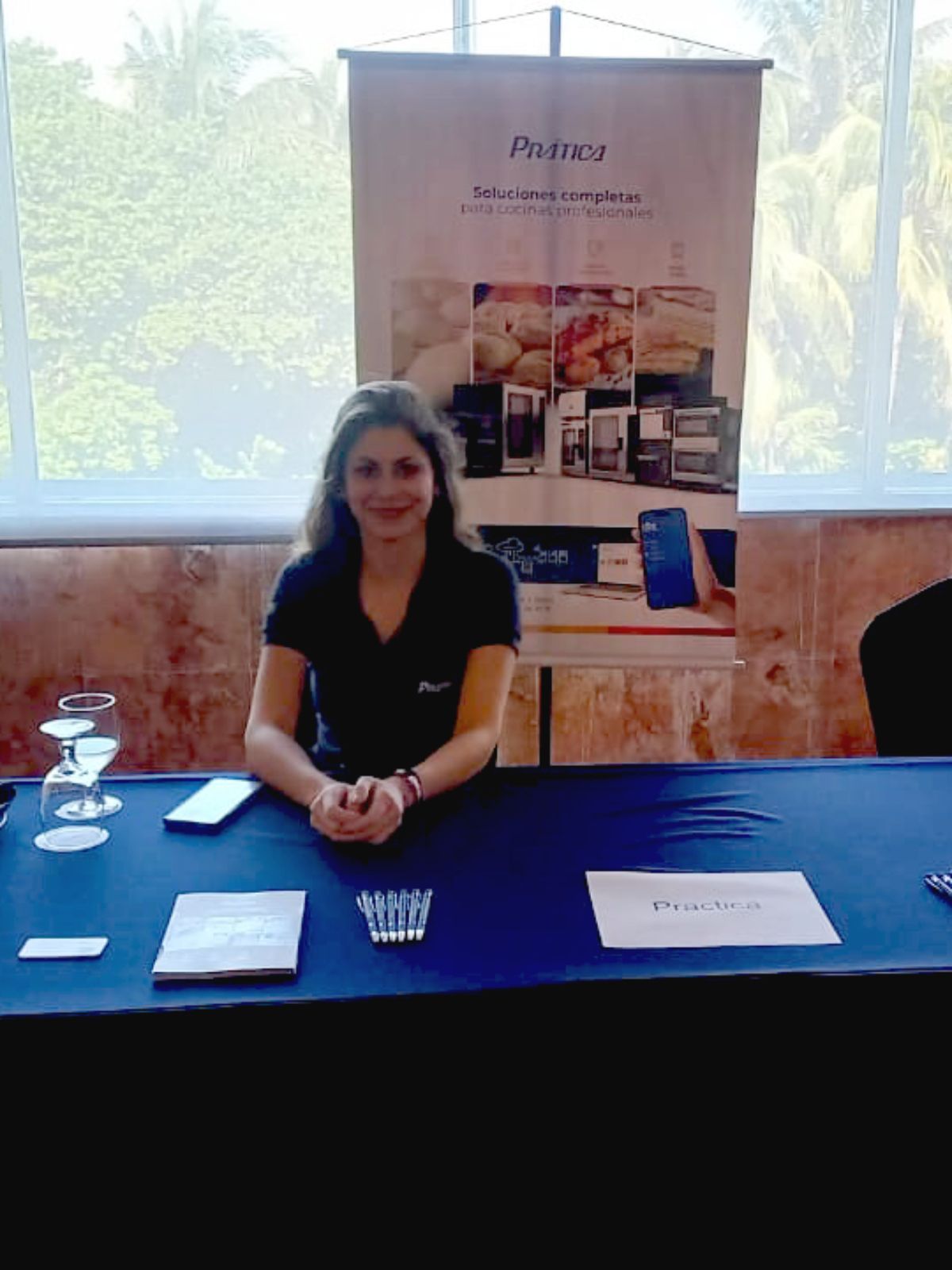 Mulher em um estande com uma faixa, sorrindo, em frente a uma janela.