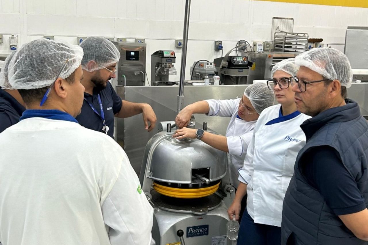 Pessoas com toucas e casacos observando uma grande máquina de processamento de alimentos de metal em uma cozinha comercial.