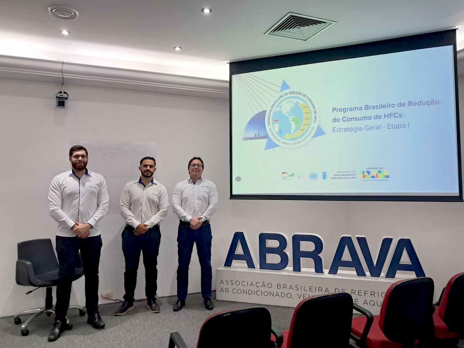 Três homens em pé em frente a uma tela com uma apresentação. Logotipo 