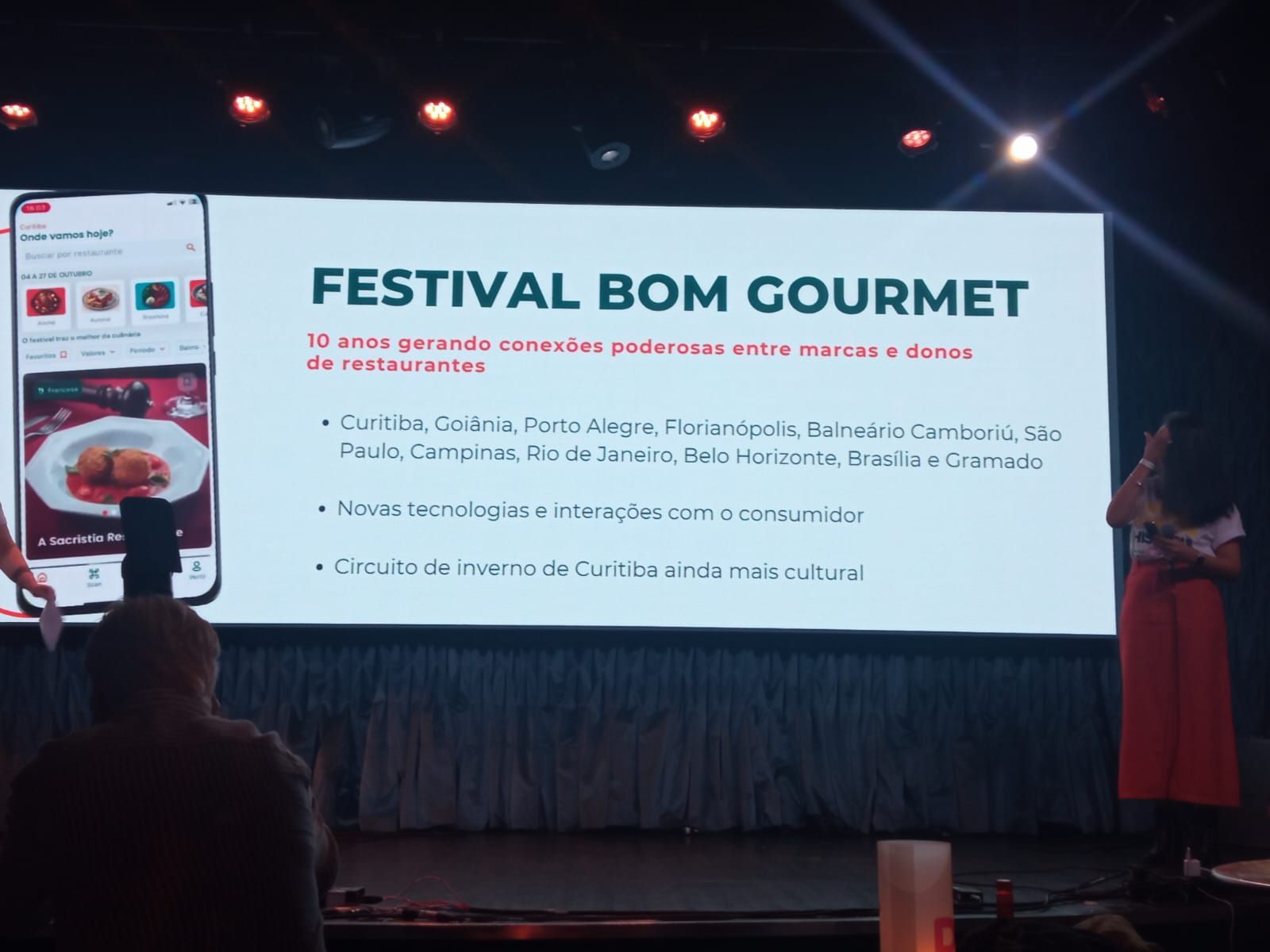 Uma mulher está parada em frente a uma tela grande que diz festival bom gourmet.