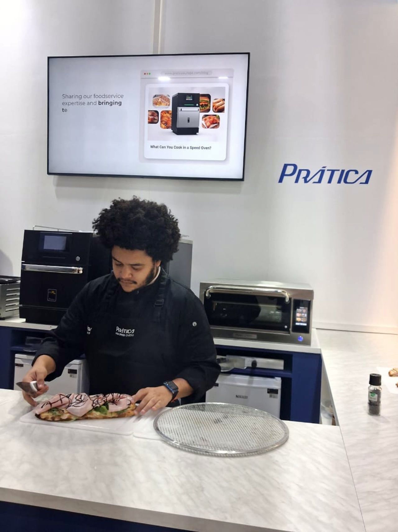 Chef preparando comida atrás de um balcão, com forno e tela de exposição. A marca 