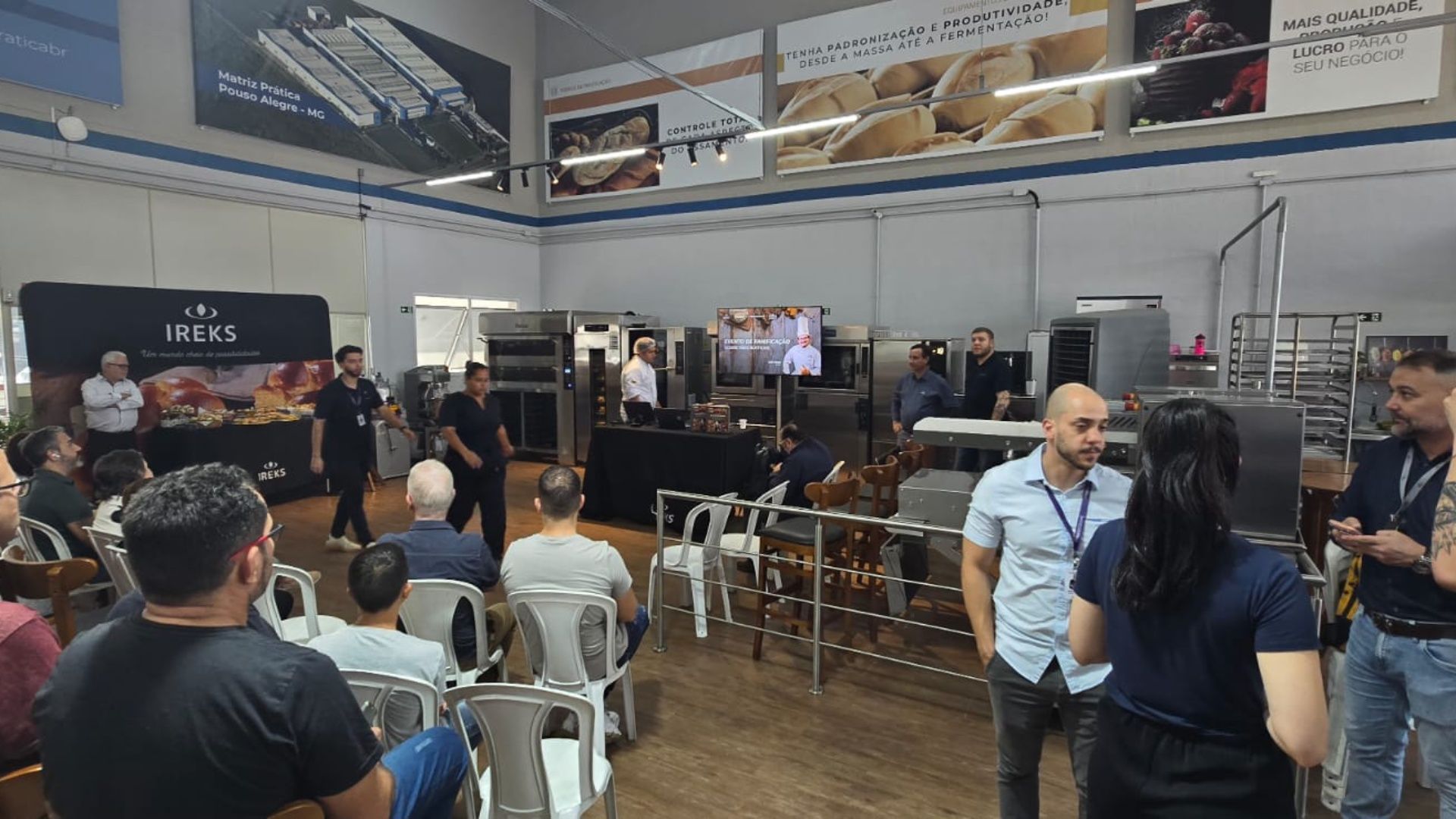 Pessoas em uma demonstração de panificação em um grande armazém. Vários fornos, exibições de pães e apresentadores são visíveis.