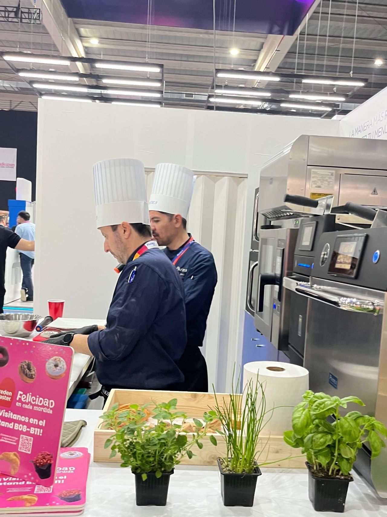 Dois chefs de chapéu branco e uniforme azul trabalham em uma cozinha comercial, com ervas em primeiro plano.
