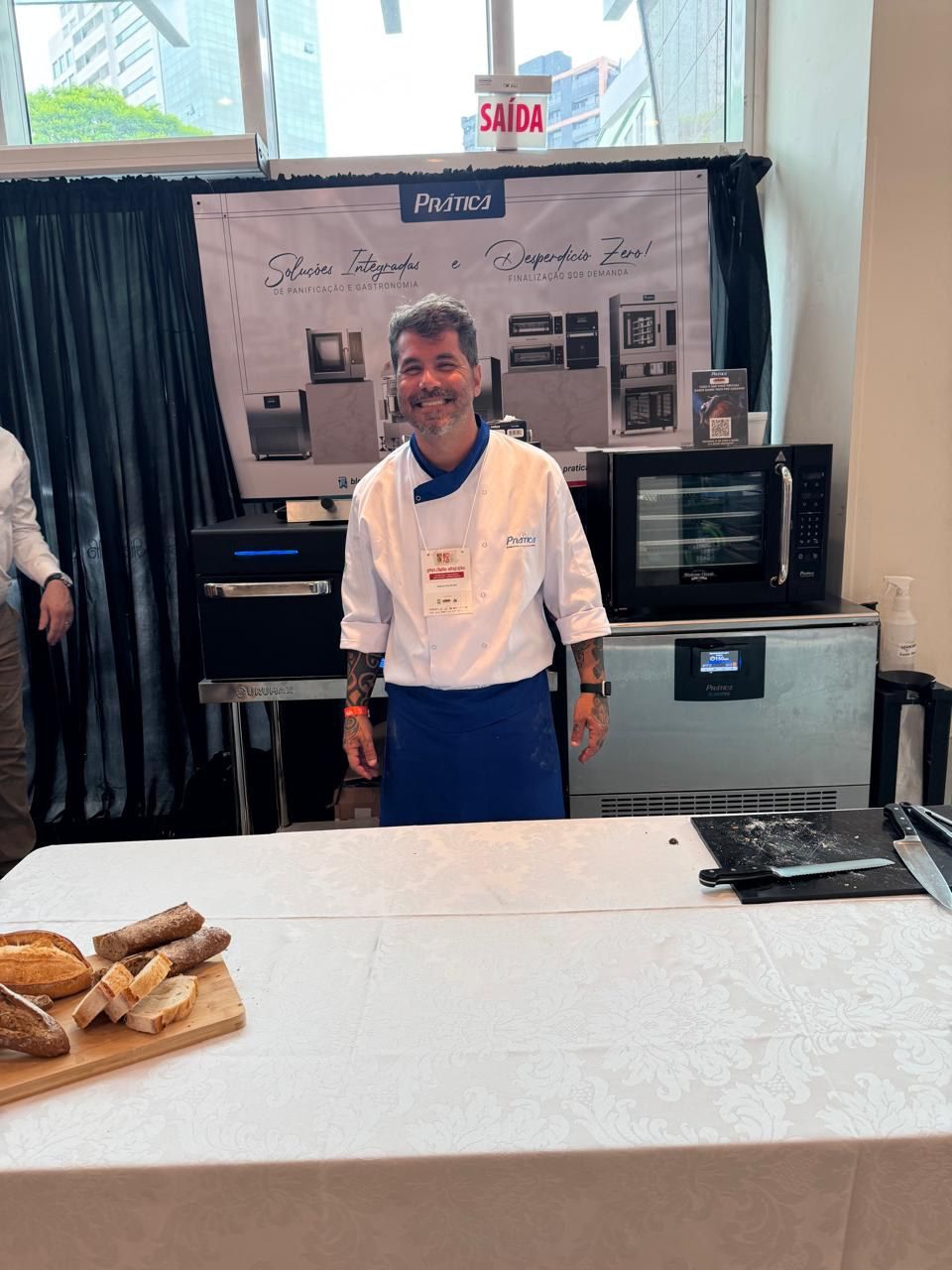 Um homem com uniforme de chef está de pé em frente a uma mesa com pão sobre ela.