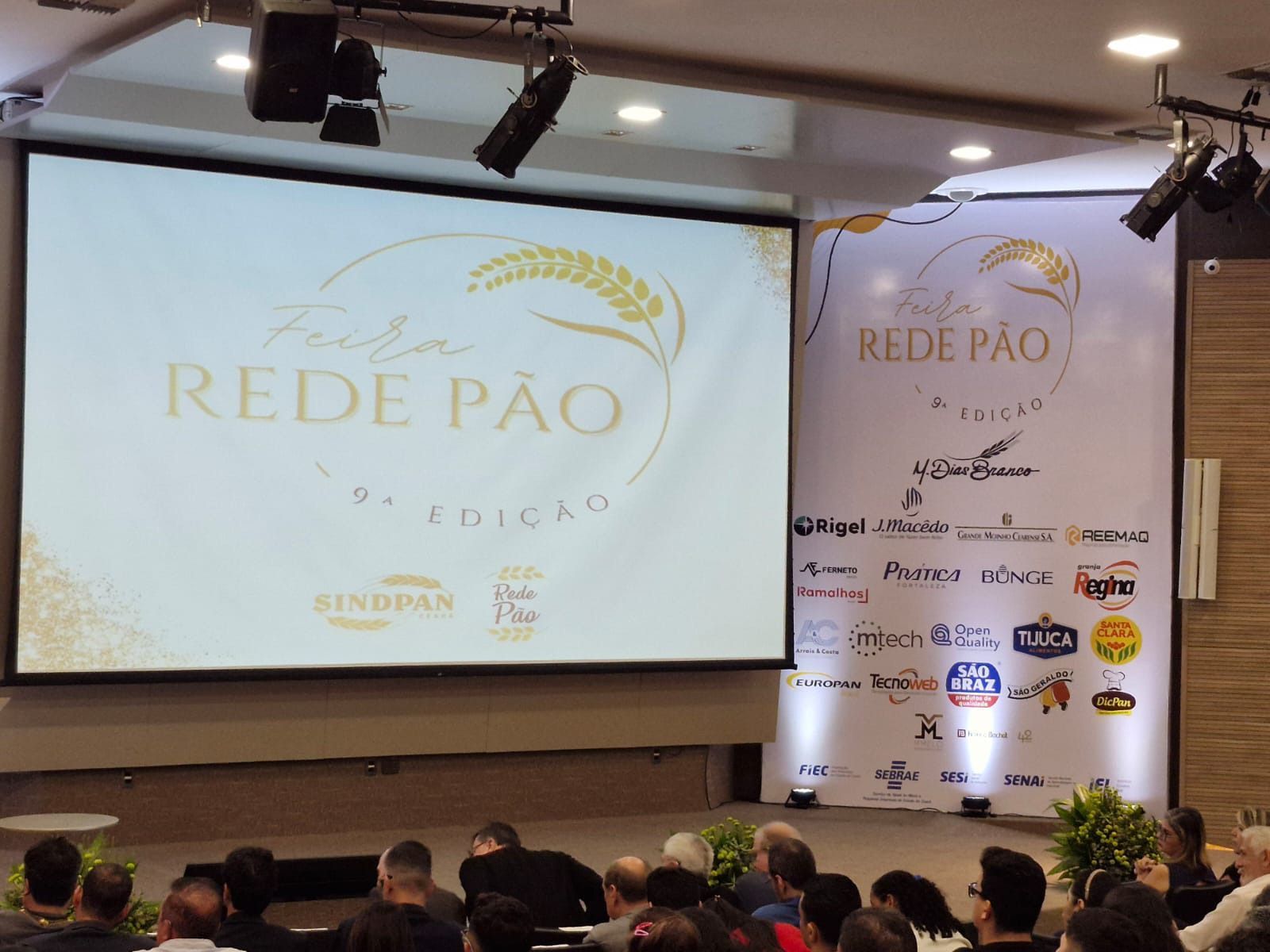 Um grupo de pessoas está sentado em frente a uma tela grande que diz rede pao