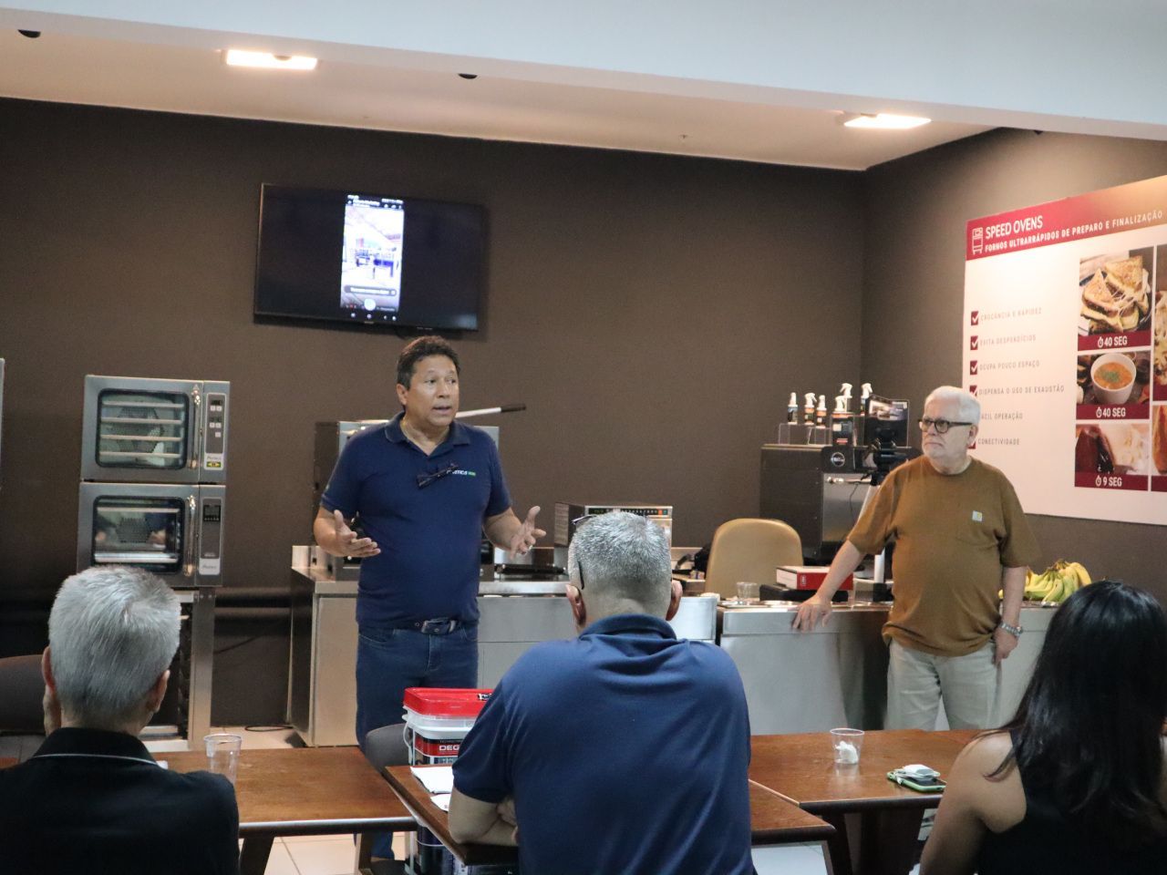 Um homem está fazendo uma apresentação para um grupo de pessoas sentadas em mesas.