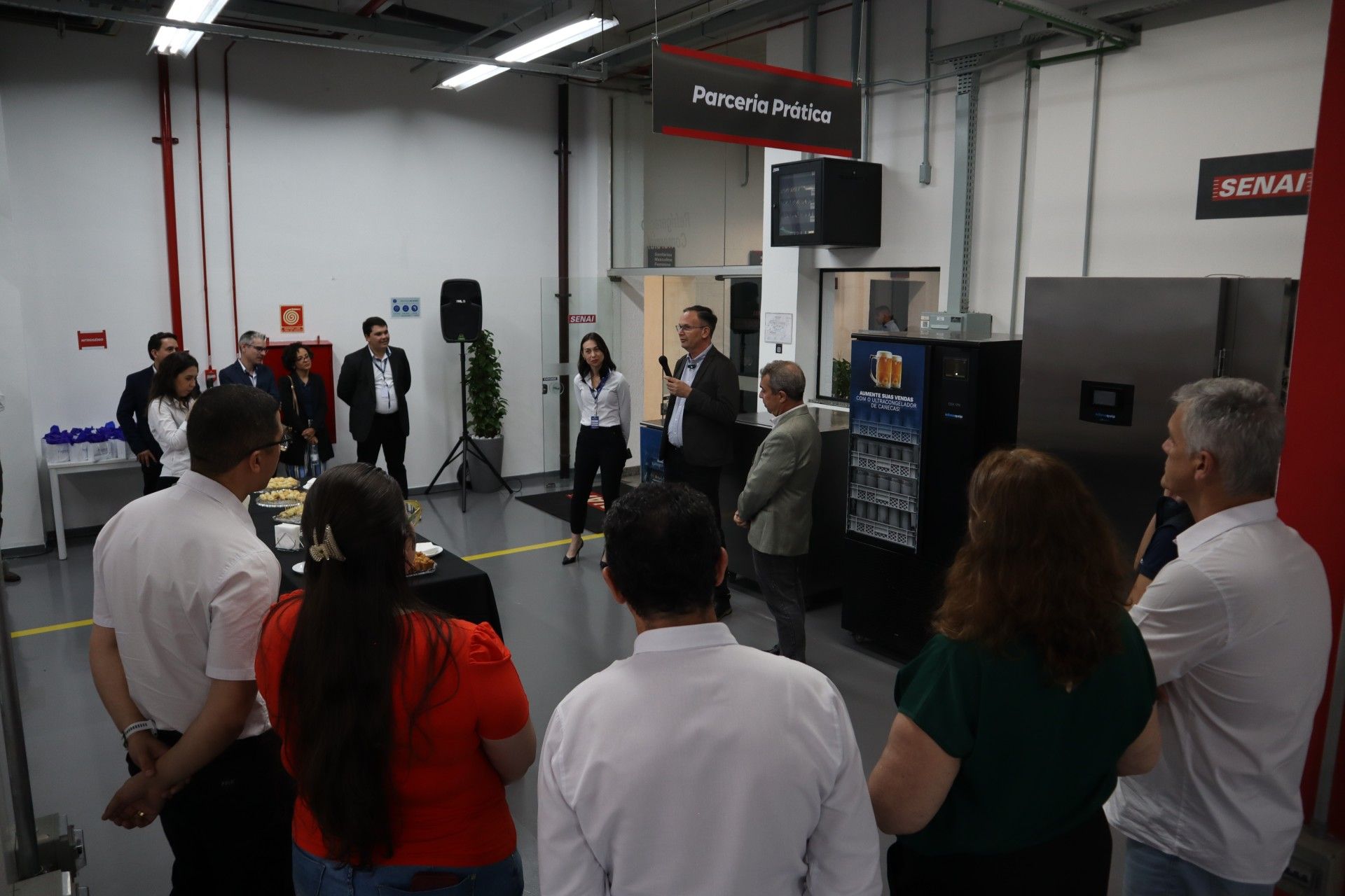 Pessoas se reúnem em um ambiente industrial bem iluminado. Duas pessoas discursam em um pódio; outras ouvem atentamente.