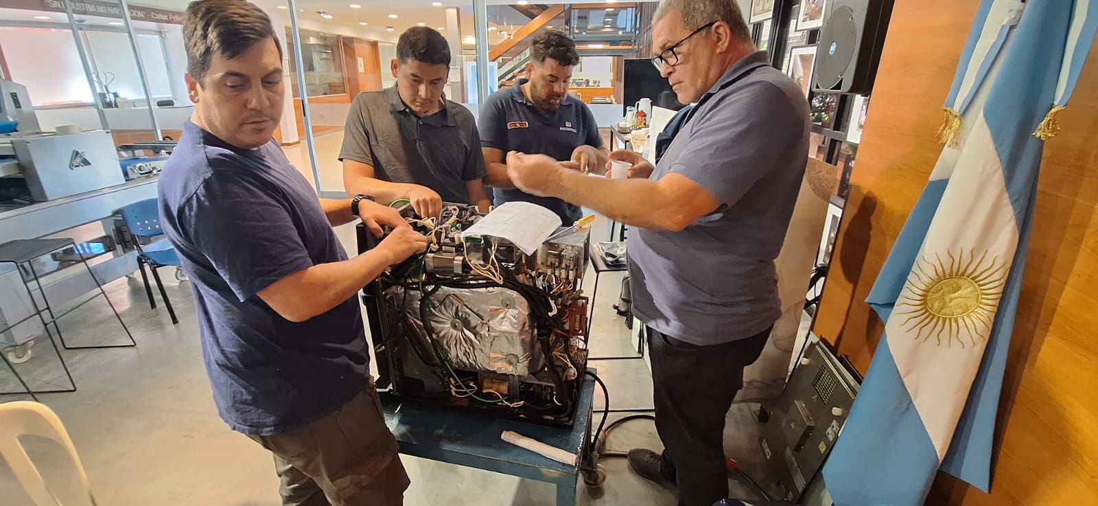 Quatro homens trabalhando em uma máquina preta, com uma bandeira argentina ao fundo.