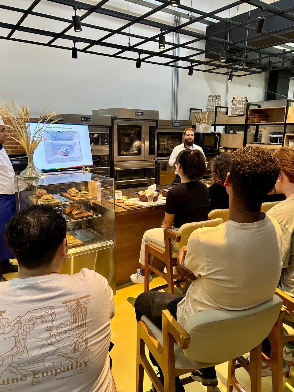 Um grupo de pessoas está sentado em cadeiras em uma padaria assistindo a uma apresentação.