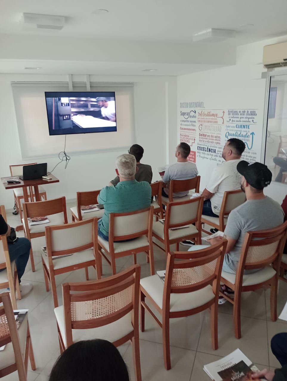 Um grupo de pessoas está sentado em cadeiras em frente a uma TV de tela plana