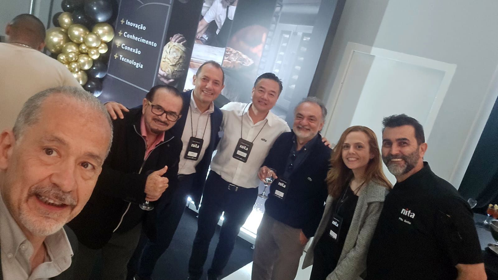 Foto de grupo em um evento: sete pessoas sorrindo, com balões ao fundo, em ambiente fechado.