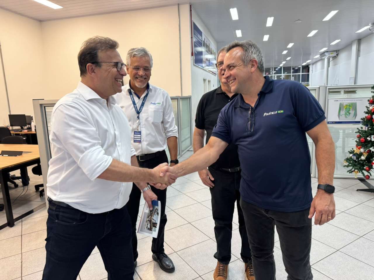 Um grupo de homens apertando as mãos em uma sala com uma árvore de natal ao fundo.