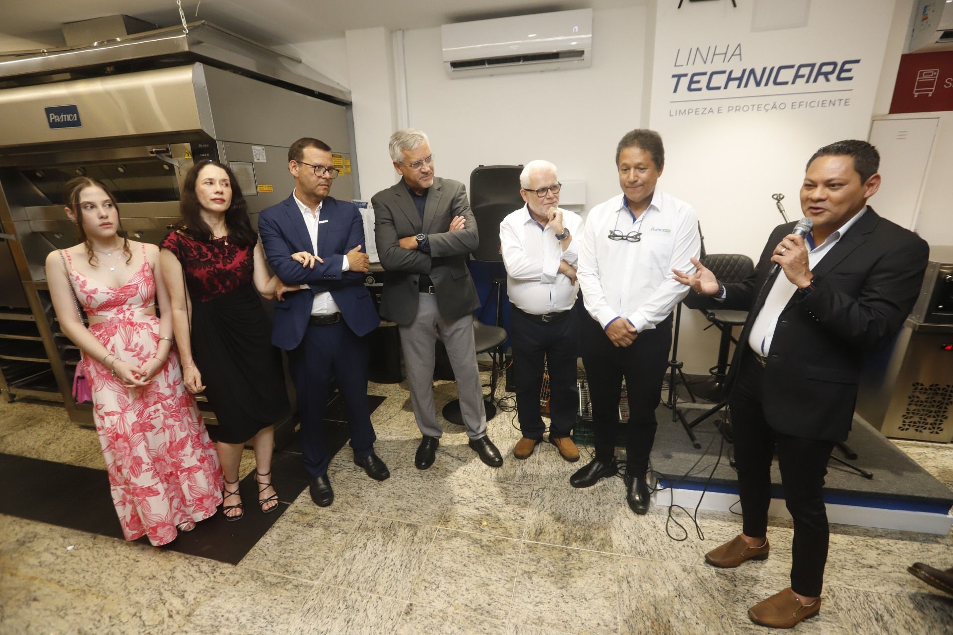 Grupo de pessoas em um evento, incluindo homens de terno e uma mulher de vestido. Um homem fala ao microfone.