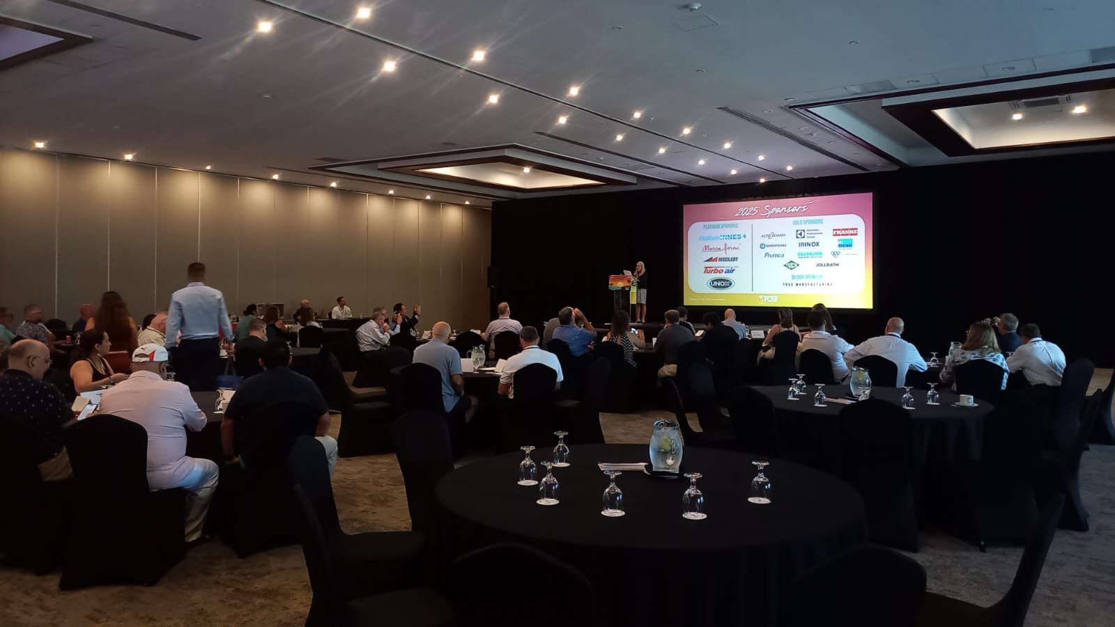 Sala de conferência com participantes sentados em mesas redondas; um apresentador fica em pé diante de uma tela exibindo uma apresentação.