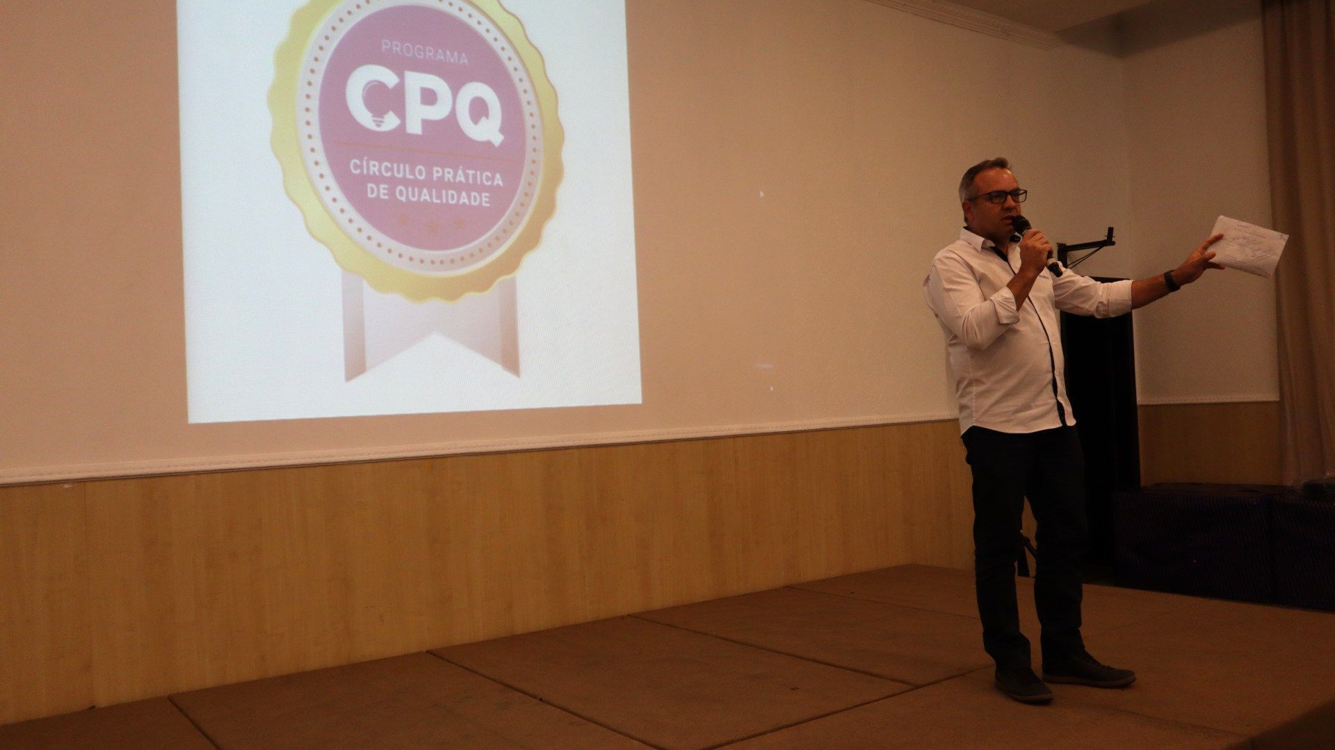 Um homem está de pé em um palco segurando um microfone em frente a uma tela de projetor.